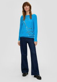 Niebieska bluzka z długim rękawem i metalicznym napisem "Piękna", w połączeniu z dżinsami w kolorze ciemnym i szerokim kroju oraz masywnymi czarnymi butami na platformie.