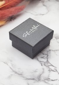 Scatola di gioielli quadrata nera con una finitura texture e un logo bianco sopra, posizionata su una superficie simile al marmo con accenti pastello morbidi nelle vicinanze.