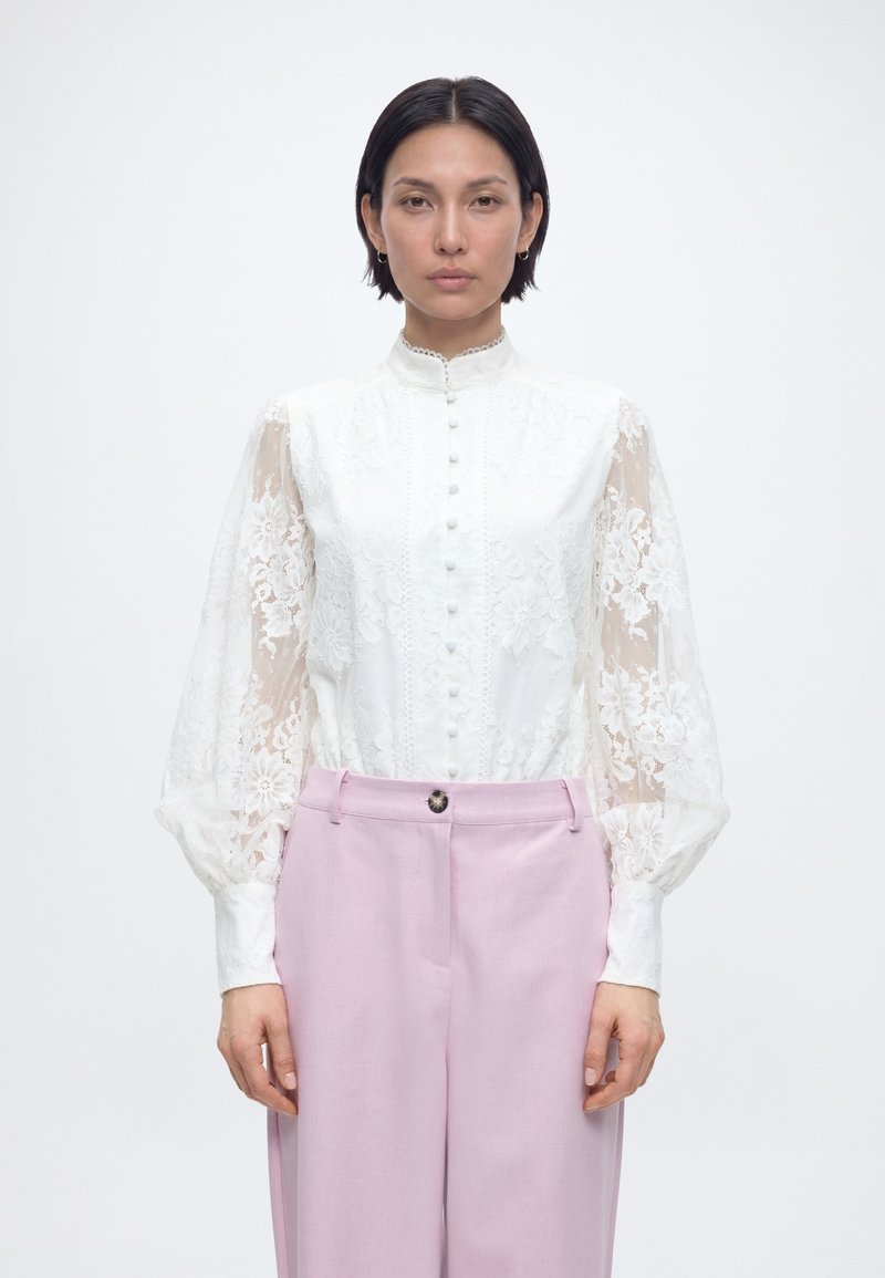 Γυναίκα με κοντά σκούρα μαλλιά που φοράει λευκό δαντελένιο μπλουζάκι με μακριά μανίκια, κουμπιά και ψηλό λαιμό, σε συνδυασμό με ανοιχτό ροζ παντελόνι.