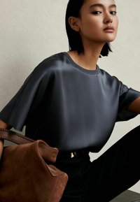 Top in satin grigio con una vestibilità ampia, maniche corte, abbinato a una borsa in suede marrone con manici intrecciati. Pantaloni neri completano l'outfit.