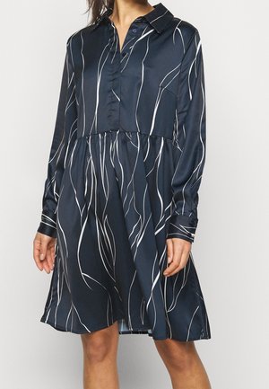 Frau trägt ein marineblaues Hemdblusenkleid mit langen Ärmeln und weißen abstrakten vertikalen Linien, steht vor einem schlichten hellen Hintergrund.
