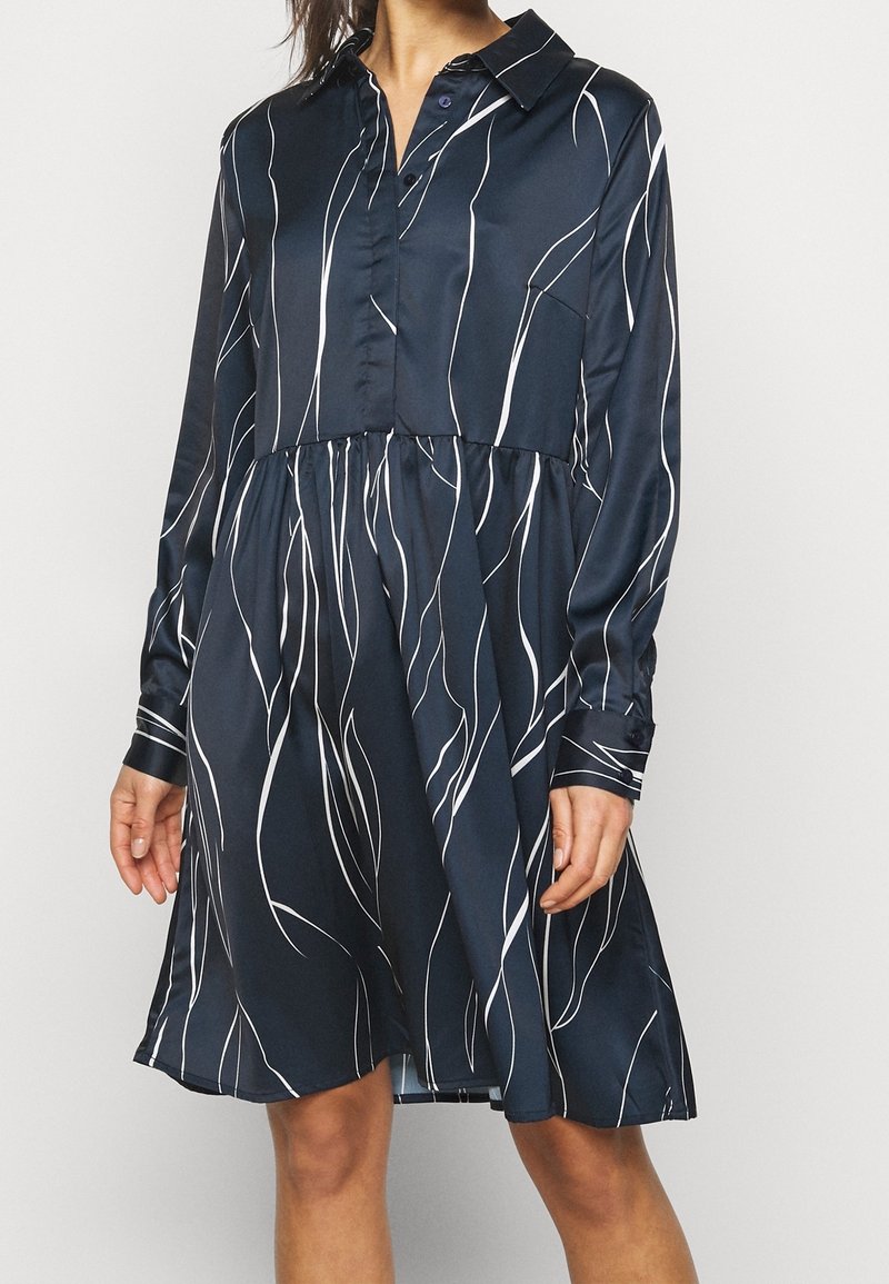 Femme portant une robe-chemise bleu marine à manches longues avec des lignes verticales abstraites blanches, debout devant un fond clair uni.