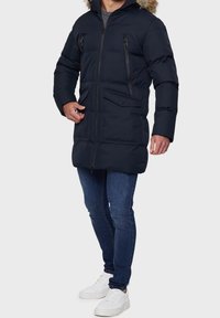 Marineblauwe donzen jas met een capuchon gevoerd met bont, ritszakken en een sluiting met knopen aan de voorkant, gecombineerd met blauwe spijkerbroek en witte sneakers.