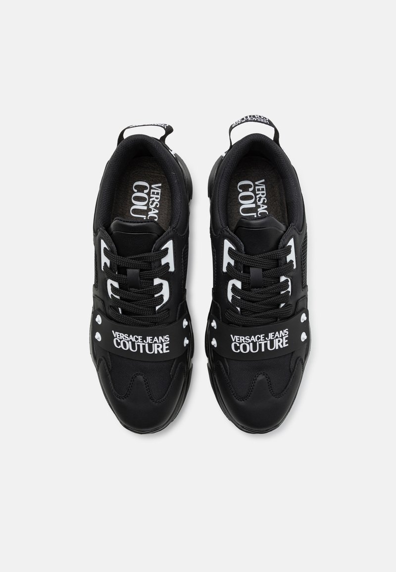 Schwarze Sneakers aus einer Kombination von glatten und strukturierten Materialien, mit weißem "VERSACE JEANS COUTURE"-Branding und markanten Akzentdetails.