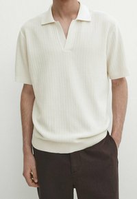 Strik polo skjorte i off-white med kort krave, ribbet tekstur og en let tætsiddende design. Kombineret med mørkebrune bukser.