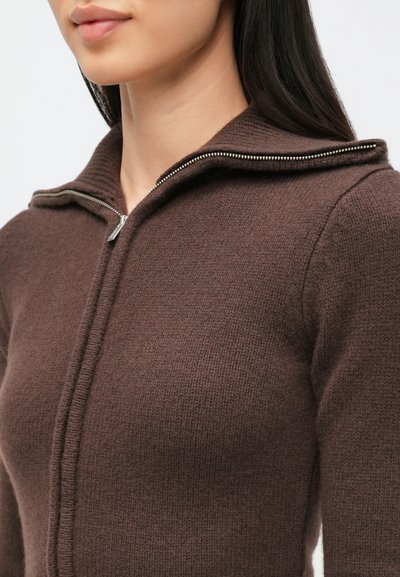 Barna cipzáros pulóver texturált felülettel, összehajtott gallérral és ezüstszínű cipzár részletekkel. Közeli kép a felsőruházatról.