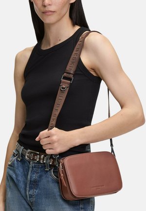 Femme portant un haut noir sans manches et un jean bleu, tenant un sac bandoulière marron avec une large bandoulière sur l'épaule.