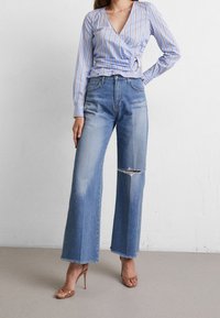 Blusa avvolgente a righe blu chiaro, jeans a vita alta e gamba larga con ginocchio strappato. Tacchi marroni con cinturini completano l'outfit.