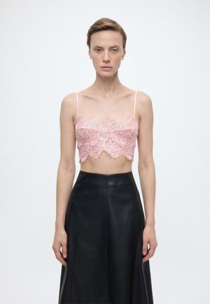 Kvinde med kort hår iført en pink blonder crop top og en sort lædernederdel med høj talje, stående foran en ensfarvet lys baggrund.