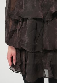 Κοντινό πλάνο ενός χεριού κοντά σε ένα στρωμένο, ημιδιαφανές σκούρο καφέ ύφασμα με διακριτή μεταλλική λάμψη και ρυτιδωτές άκρες.