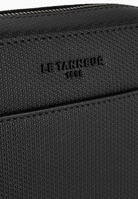 Bolso de cuero negro texturizado con el logo en relieve "LE TANNEUR 1898", que presenta un cierre de cremallera suave y un diseño geométrico limpio.