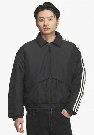 Giovane uomo con capelli neri lisci che indossa una giacca nera con cerniera e strisce bianche sulle maniche e pantaloni neri, in piedi e rivolto verso di fronte.