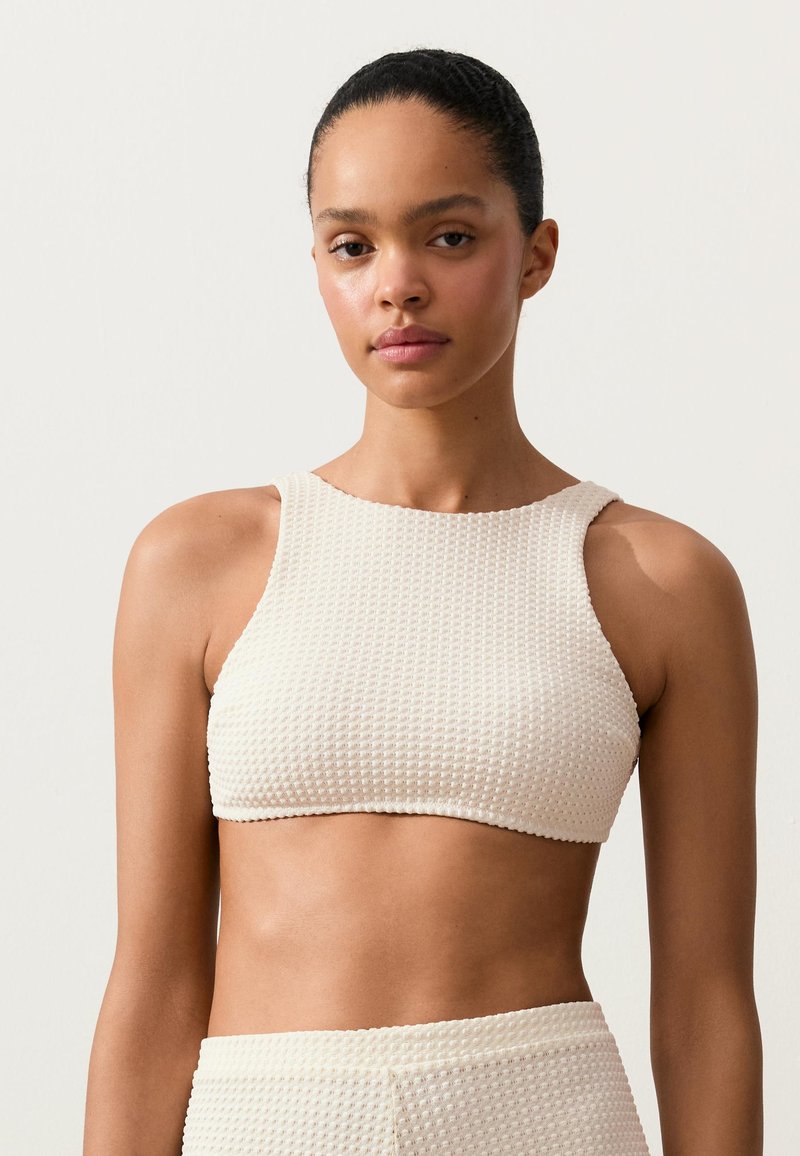 Jeune femme aux cheveux foncés attachés vers l’arrière, portant un crop top blanc texturé sans manches et un bas taille haute assorti sur fond uni.
