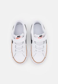 White Nike sneakers with black accents, featuring a strap, lace closure, and brown rubber outsole. Textured fabric with mesh lining.