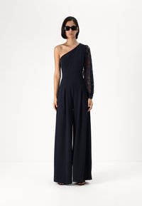 Navy jumpsuit featuring one long lace sleeve, a fitted bodice, wide legs, and a structured design. Worn with dark sunglasses.