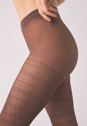 Közeli kép egy személyről, aki barna mintás átlátszó harisnyát visel, kezét a derekára téve, semleges háttér előtt.