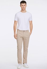 Gaiši bēšas krāsas bikses chinos stila, kombinētas ar vienkāršu baltu T-kreklu un baltām sporta kurpēm. Bikšu audums ir gluds, un griezums pieguļošs.