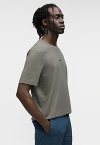 Κοντομάνικο, ελαφρώς πράσινο t-shirt από μαλακό βαμβάκι με στρογγυλή λαιμόκοψη· διαθέτει μικρό μαύρο λογότυπο στο στήθος. Συνδυάζεται με μπλε παντελόνι.