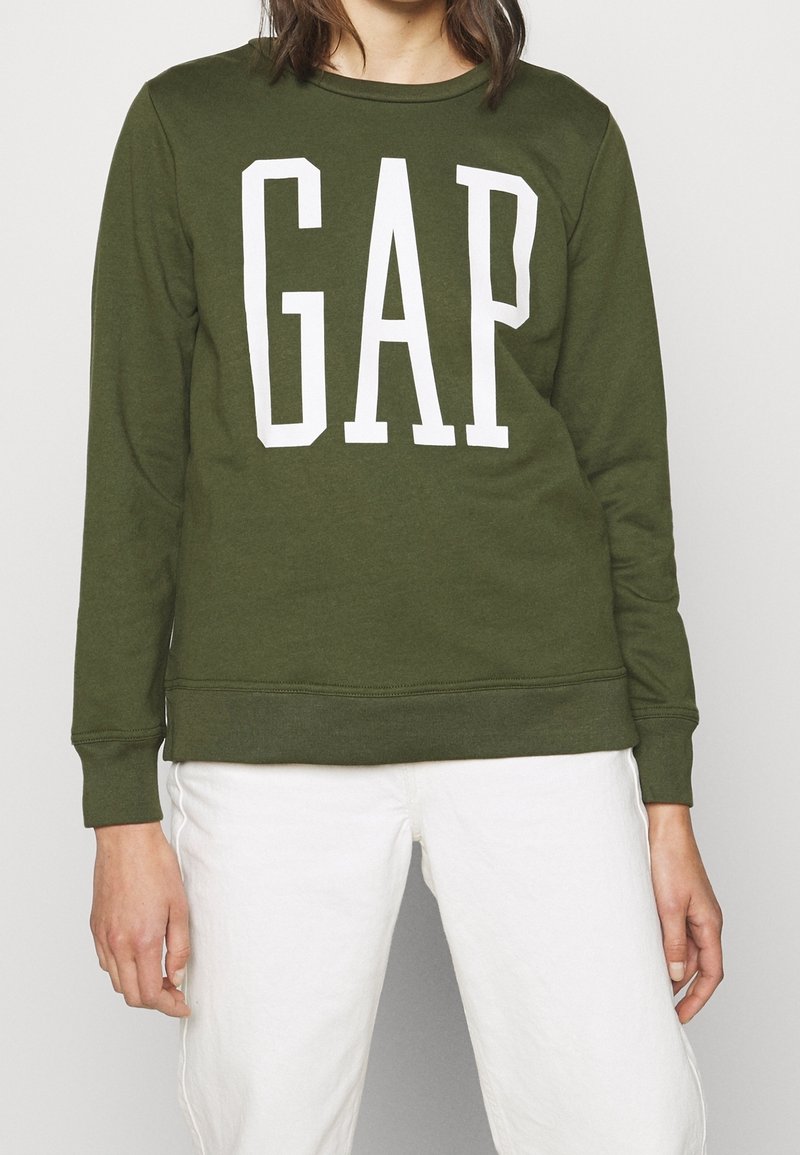 Personne portant un sweat-shirt à manches longues vert olive avec de grandes lettres blanches "GAP" et un pantalon blanc sur un fond uni.