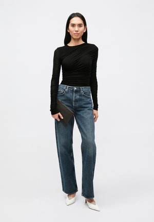 Zwarte top met lange mouwen van gestructureerde stof, hoge halslijn; lichtblauwe jeans met hoge taille; witte puntige hakken; houdt een kleine zwarte clutch vast.