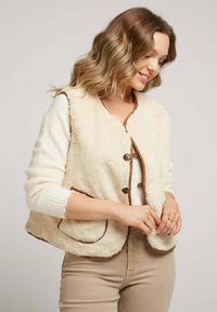 Femme aux cheveux ondulés en train de boutonner un gilet en peluche crème avec des bordures brunes sur un léger pull, portant un pantalon beige, sur un fond uni.