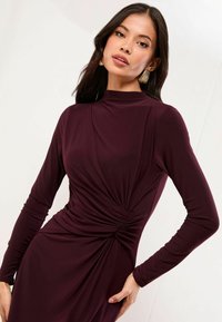 Rochie burgundy cu mânecă lungă, cu un gât înalt, având detalii plisate la talie și un material neted, cu textură.