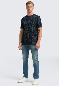 Man wearing a dark patterned short-sleeve t-shirt, faded blue jeans, and white sneakers standing against a plain background.