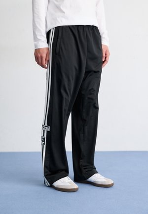 Mężczyzna w czarnych spodniach dresowych Adidas z białymi paskami i białych tenisówkach stoi na niebieskiej podłodze na tle białej ściany.