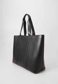 Sac tote noir en cuir lisse, avec de longues poignées plates et un logo en relief sur les côtés. Design structuré et minimaliste.