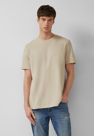 Jeune homme aux cheveux courts et bruns portant un t-shirt beige à manches courtes et un jean bleu, debout devant un fond gris uni.