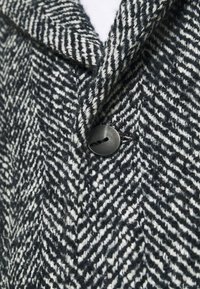 Cappotto con motivo pied de poule in tweed bianco e nero, caratterizzato da un colletto a rever e una chiusura con bottone rotondo nero. Dettaglio del tessuto strutturato visibile.