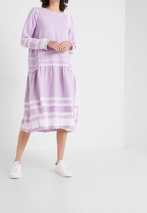 Femme portant une robe midi à manches longues à motifs lavande et blanche avec une jupe à volants et des baskets blanches, debout sur un sol blanc.
