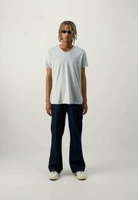 Ανοιχτό γκρι T-shirt με V-λαιμόκοψη, χαλαρές ναυτικές παντελόνες και λευκά sneakers. Μοντέλο με γυαλιά ηλίου και πλεξούδα στέκεται μπροστά σε απλό φόντο.