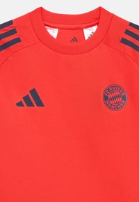 Κόκκινο αθλητικό μπλουζάκι από λεία ύφανση υφάσματος. Διαθέτει λογότυπο Adidas σε ναυτικό μπλε χρώμα στο αριστερό στήθος και τρεις ρίγες στους ώμους. Εμφανίζει το έμβλημα της FC Bayern München.