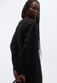 Μαύρο φόρεμα φούτερ με στρογγυλή λαιμόκοψη, φαρδιά μανίκια και ορατό φερμουάρ που κατεβαίνει στην πλάτη. Ύφασμα με λείο υφή.