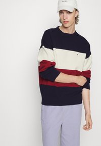 Maglione lavorato a maglia con strisce orizzontali in blu navy, crema e rosso. Maniche lunghe con polsini a costine. Indossato con pantaloni chiari.