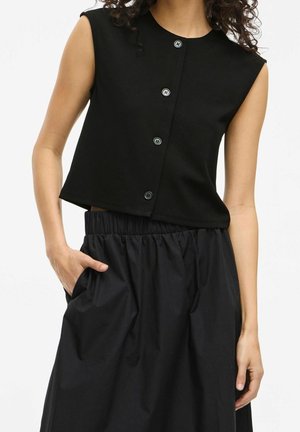 Vrouw draagt een mouwloze zwarte top met knopen en een zwarte rok met hoge taille en elastische tailleband, met één hand in de zak, tegen een witte achtergrond.