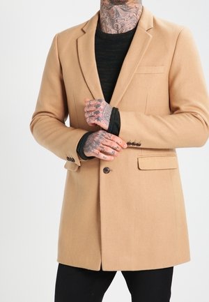 Homme avec des tatouages au cou et à la main, portant un blazer beige sur un haut noir à manches longues, ajustant le poignet gauche avec la main droite.