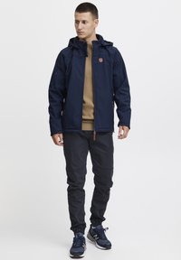 Veste imperméable bleu marine avec capuche, dotée d'une fermeture éclair, de poches latérales et d'un écusson logo marron. Pantalon sombre et chaussures de sport.