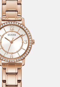 Roosa kuldne metallkell ümmarguse käekellaga, kristallaktsentide ümber bezelil ning valge kellaga, mis sisaldab kuldseid tunni markereid ja käsi.