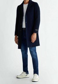 Mörkblå ullkappa med slagkrage och tre knappar, i kombination med mörkblå jeans och vita sneakers. Har en ficketikett.