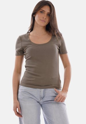 Giovane donna con lunghi capelli castani che indossa una maglietta verde oliva a maniche corte e jeans chiari, in piedi contro uno sfondo uniforme.