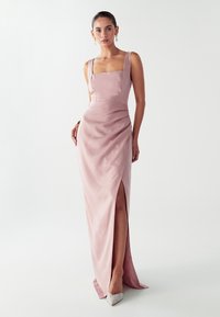 Moteris, vilkinti be rankovių, kvadratinio iškirpimo, dulkėto rožinio satino suknelę su aukšta iškirpte ir blizgančiais smailiais kulniukais, prieš paprastą baltą foną.