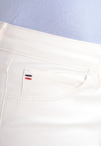 Jeans bianchi con una texture liscia, caratterizzati da una piccola tasca anteriore con un dettaglio di cucitura a righe rosse, bianche e blu.