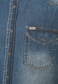 Camicia di denim blu medio con zone sbiadite, dotata di bottoni a scatto argentati, una tasca sul petto e etichetta LTB sulla tasca.