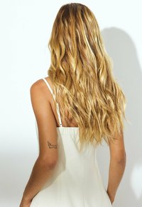 Vit klänning med tunna axelband, med en slank design. Långt, vågigt blont hår och en liten tatuering på armen.