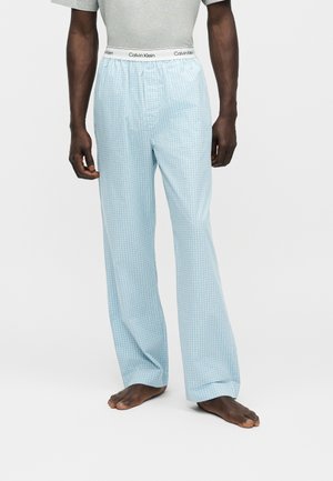 Hombre con pantalones de pijama de cuadros azul claro con banda elástica con el logo de Calvin Klein, de pie descalzo sobre fondo blanco.