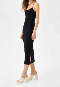 Schwarzes Midikleid mit strukturiertem Muster, dünnen Trägern und Seitenschlitzen. Das Modell kombiniert es mit beigen Plateauschuhen mit glänzender Oberfläche.