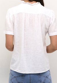 Blusa bianca in lino con maniche corte, tessuto arricciato sul retro e dettaglio di scollatura a volant. Abbinata a jeans in denim blu.