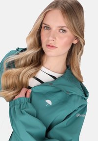 Türkizkék vízálló dzseki kapucnival, rugalmas mandzsettákkal és fehér esernyőlogóval. Csíkozott pulóver fölött viselve, laza stílust tükröz.